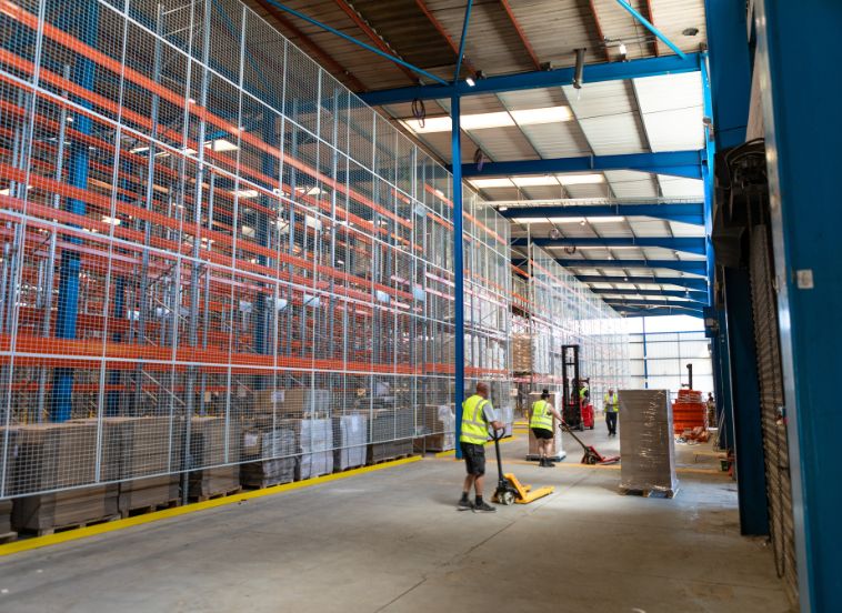 Inside warehouse with floor to ceiling racking with people in yellow hi-vis working with pump trucks